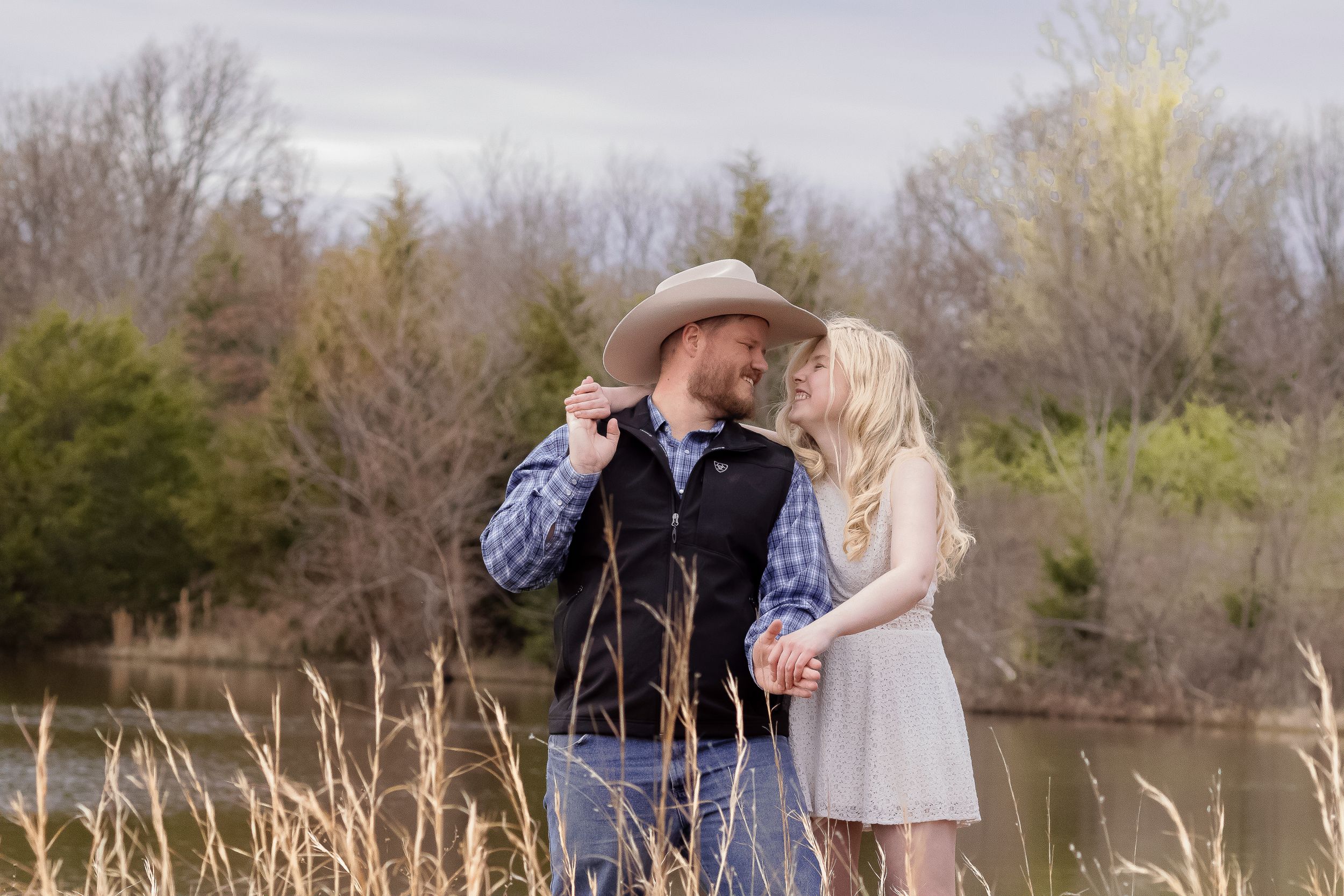 Couple sharing romantic moment in field near lake with man wearing cowboy hat.