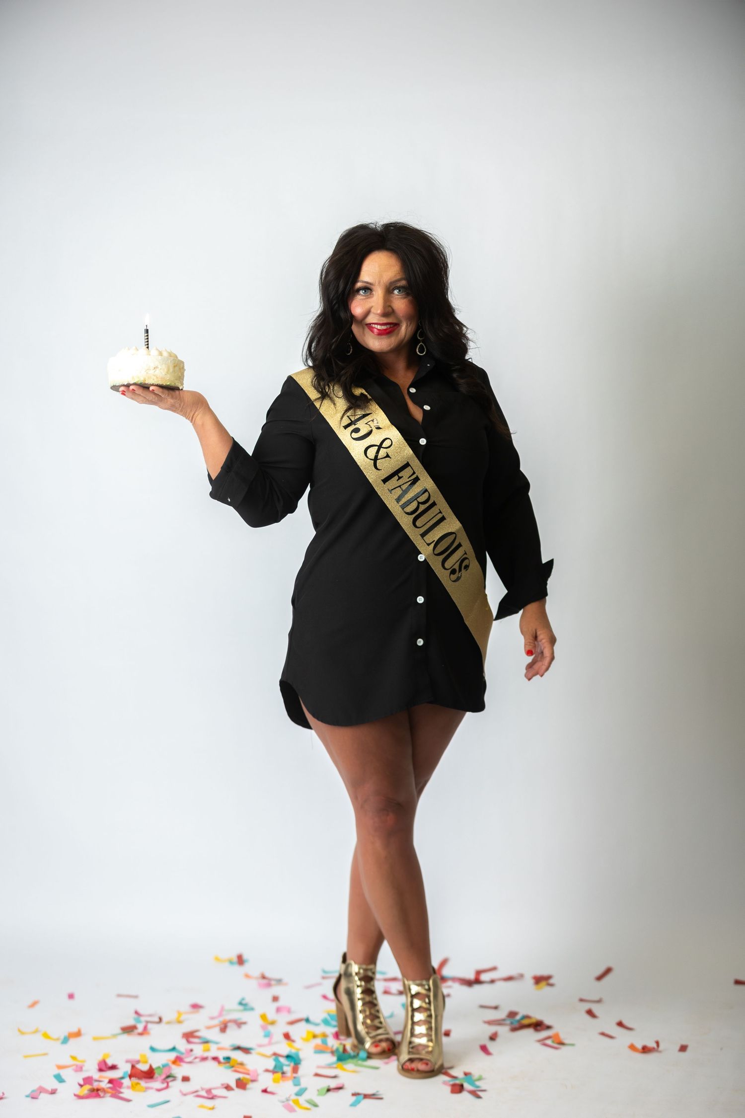 Woman in black blazer wearing gold sash celebrates with colorful confetti on white background.