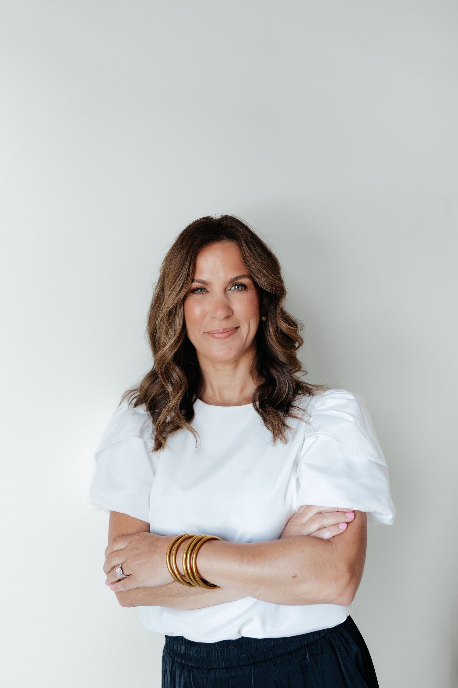 Professional portrait of a person in a white puff sleeve top and dark pants standing against a light gray wall with stacked bracelets.