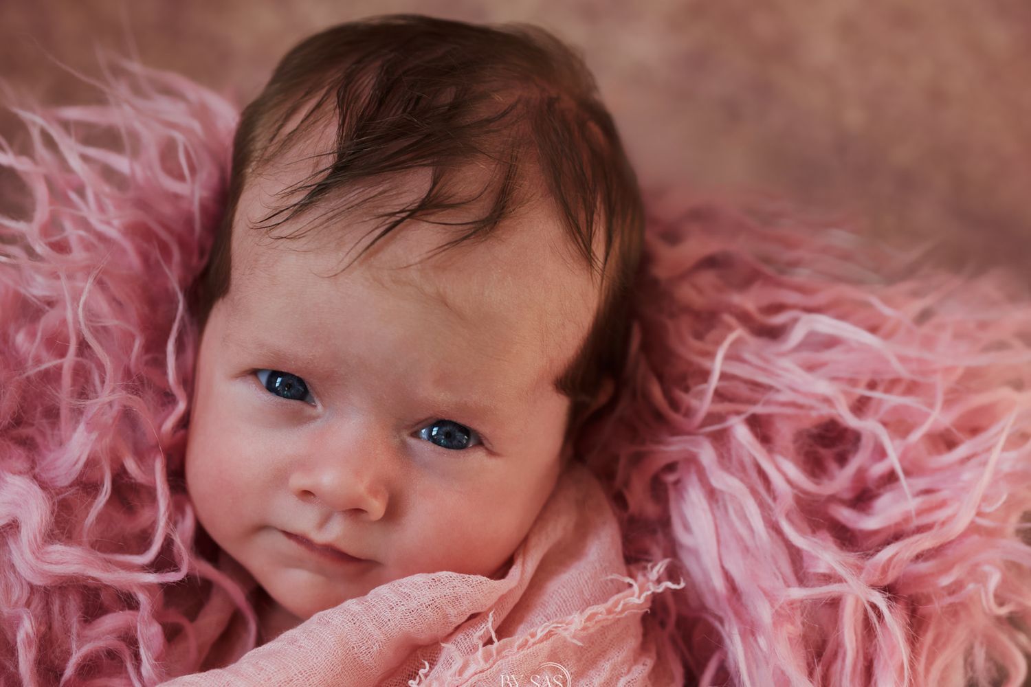 Newborn baby girl with blue eyes smiling in pink fluffy blanket wrap for professional newborn photography session.