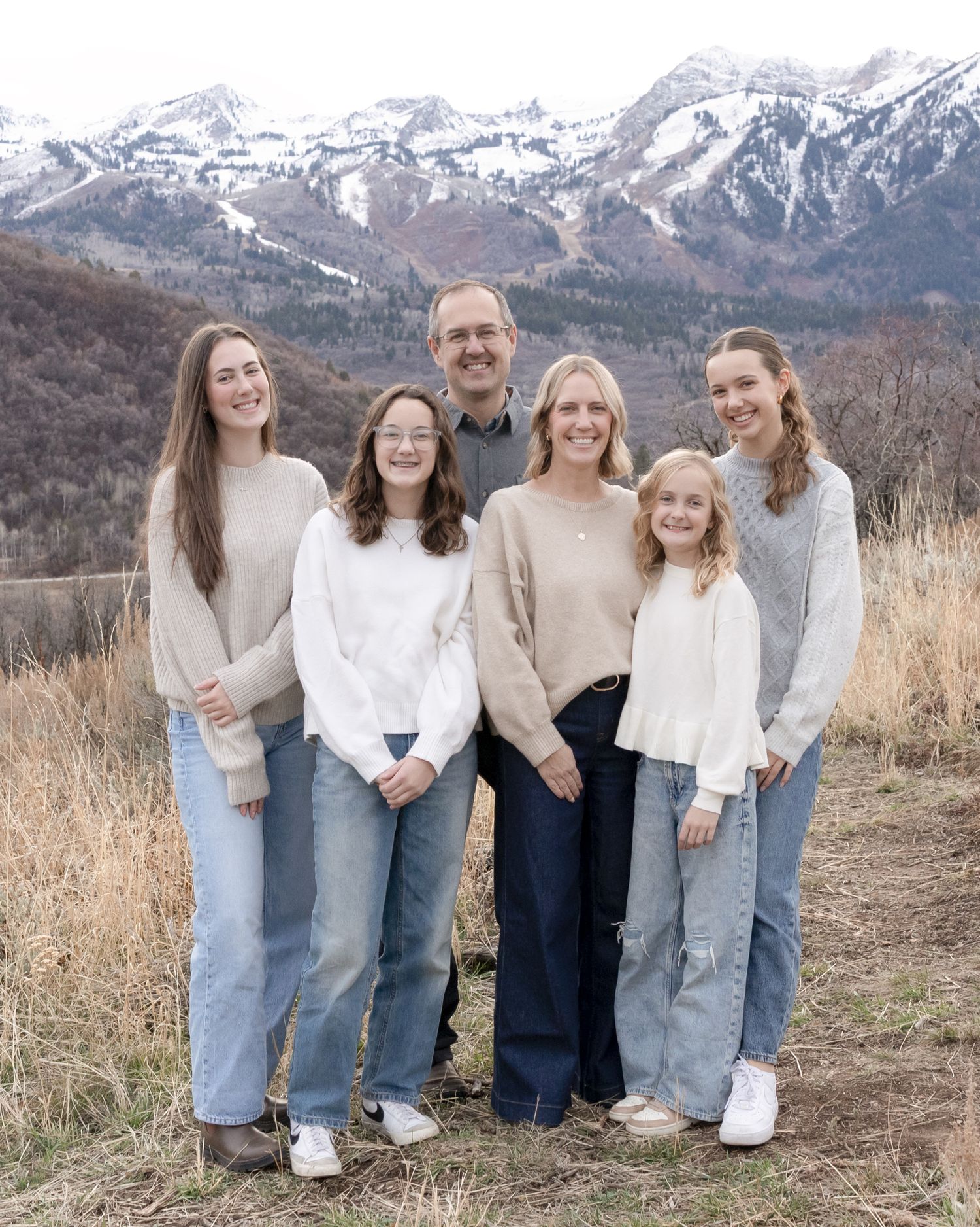 A family group poses together on a hillside with snow-capped mountains in the background wearing coordinated white and neutral outfits.