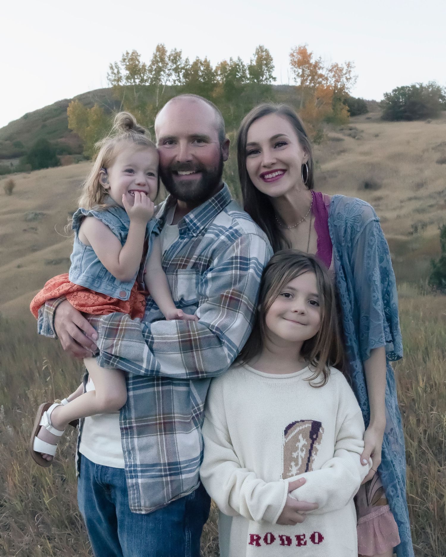 A family poses together outdoors in a grassy field with rolling hills and autumn trees in the background.
