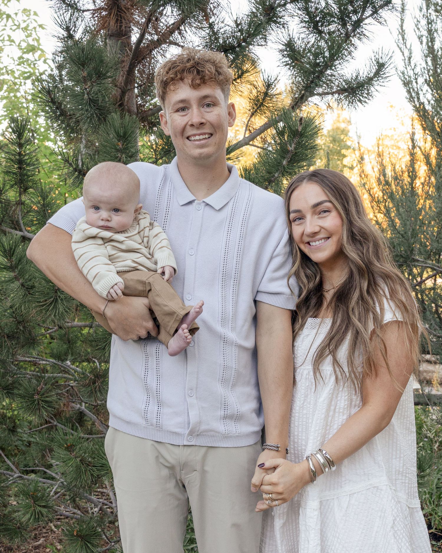 A family wearing coordinated white and beige outfits poses together in front of evergreen trees during a sunset photoshoot.