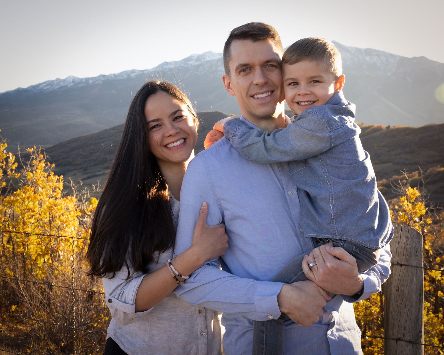 Family poses together outdoors during golden hour with mountains and autumn foliage in background.