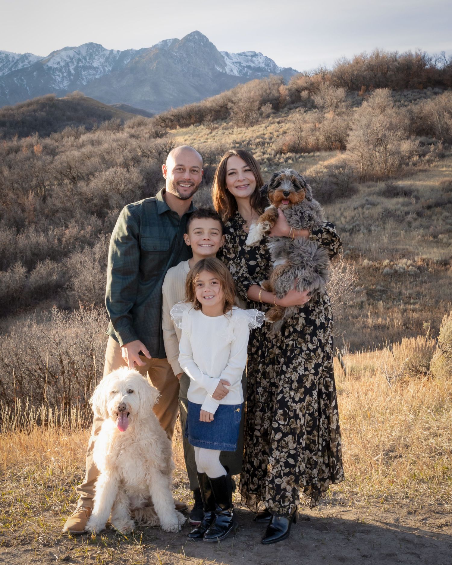 A family poses together with their fluffy white dog against a mountainous autumn landscape backdrop.