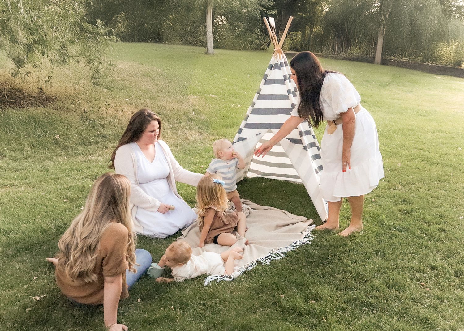 Mothers and young children sitting on a blanket by a stripe teepee during a relaxed outdoor family photo shoot in Ogden Valley Utah