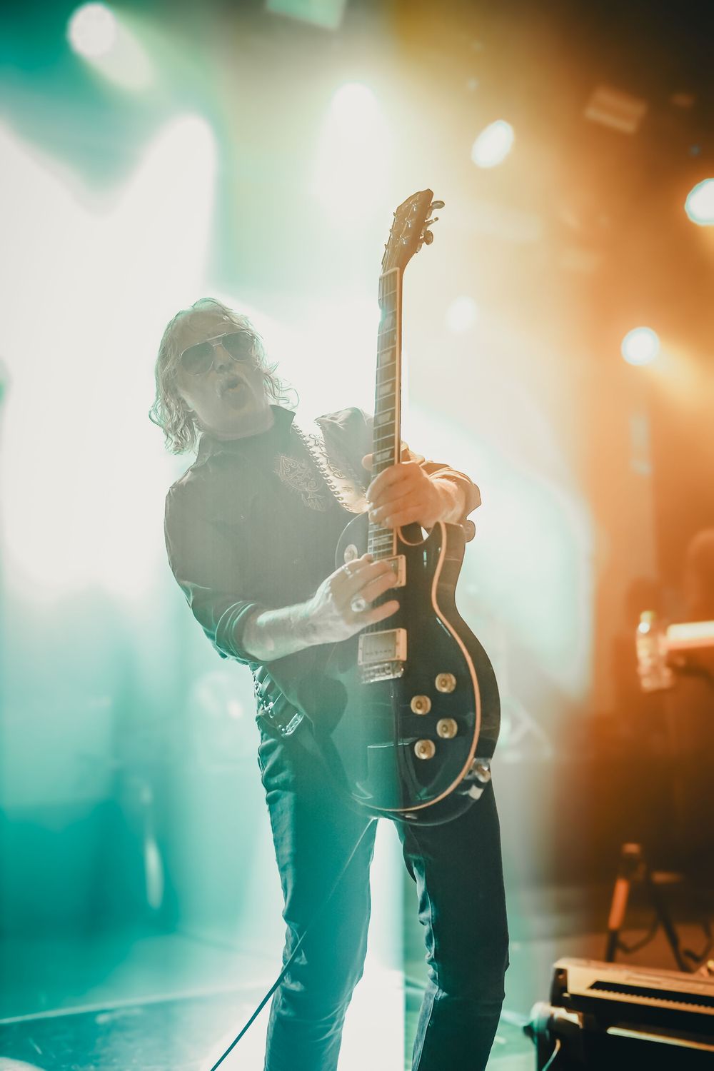 Guitarist performs on stage with dramatic teal and orange lighting creating atmospheric silhouette effect.