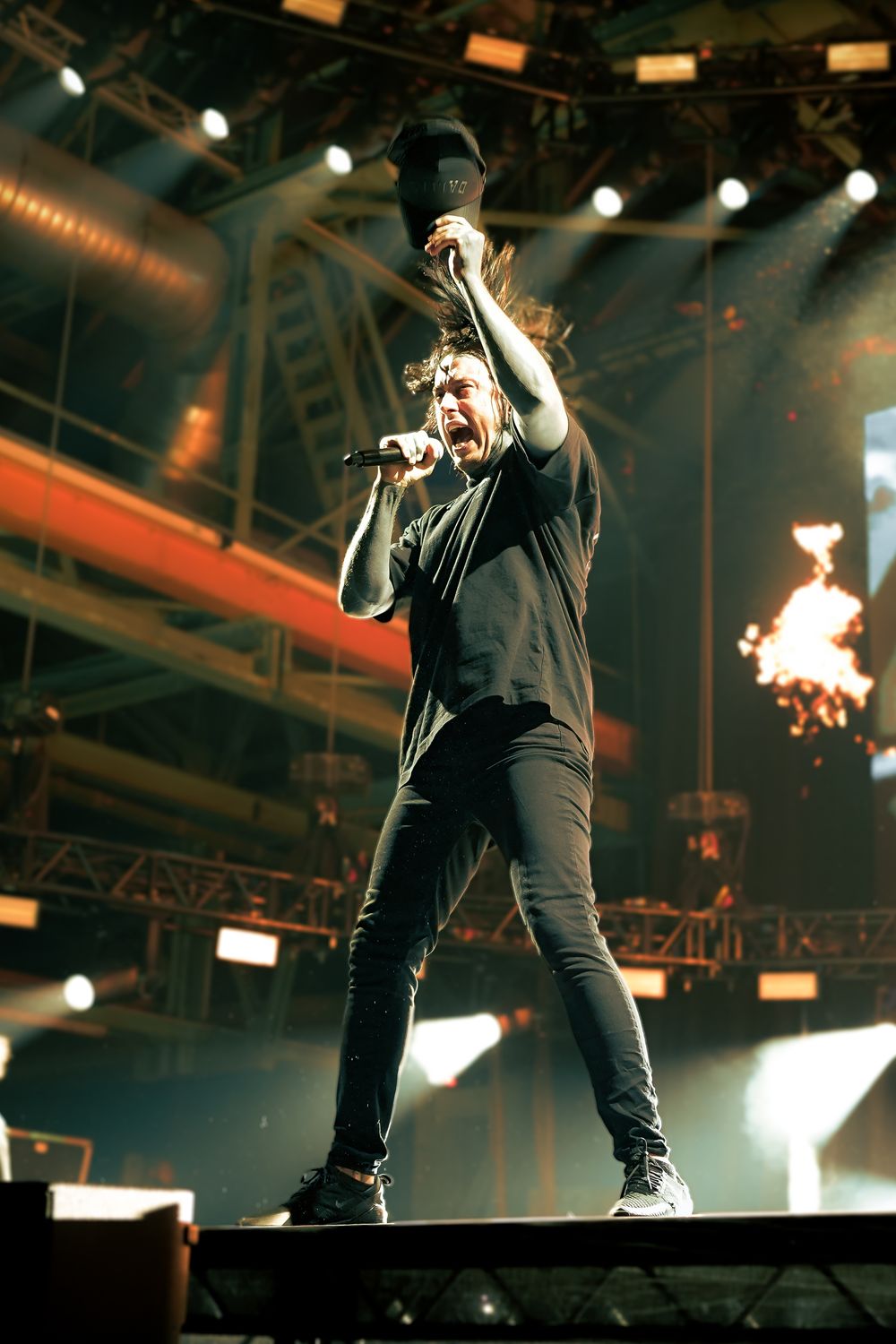 Performer in black outfit energetically singing on stage with dramatic lighting and pyrotechnic effects in background.