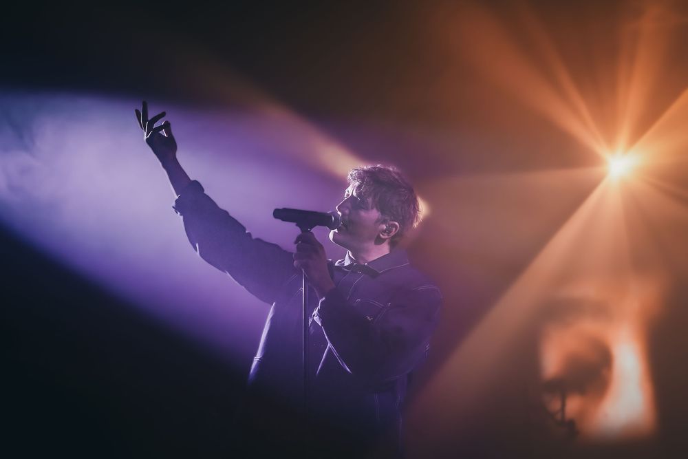 A silhouetted performer on stage raises their hand dramatically against purple and orange spotlights during a show.