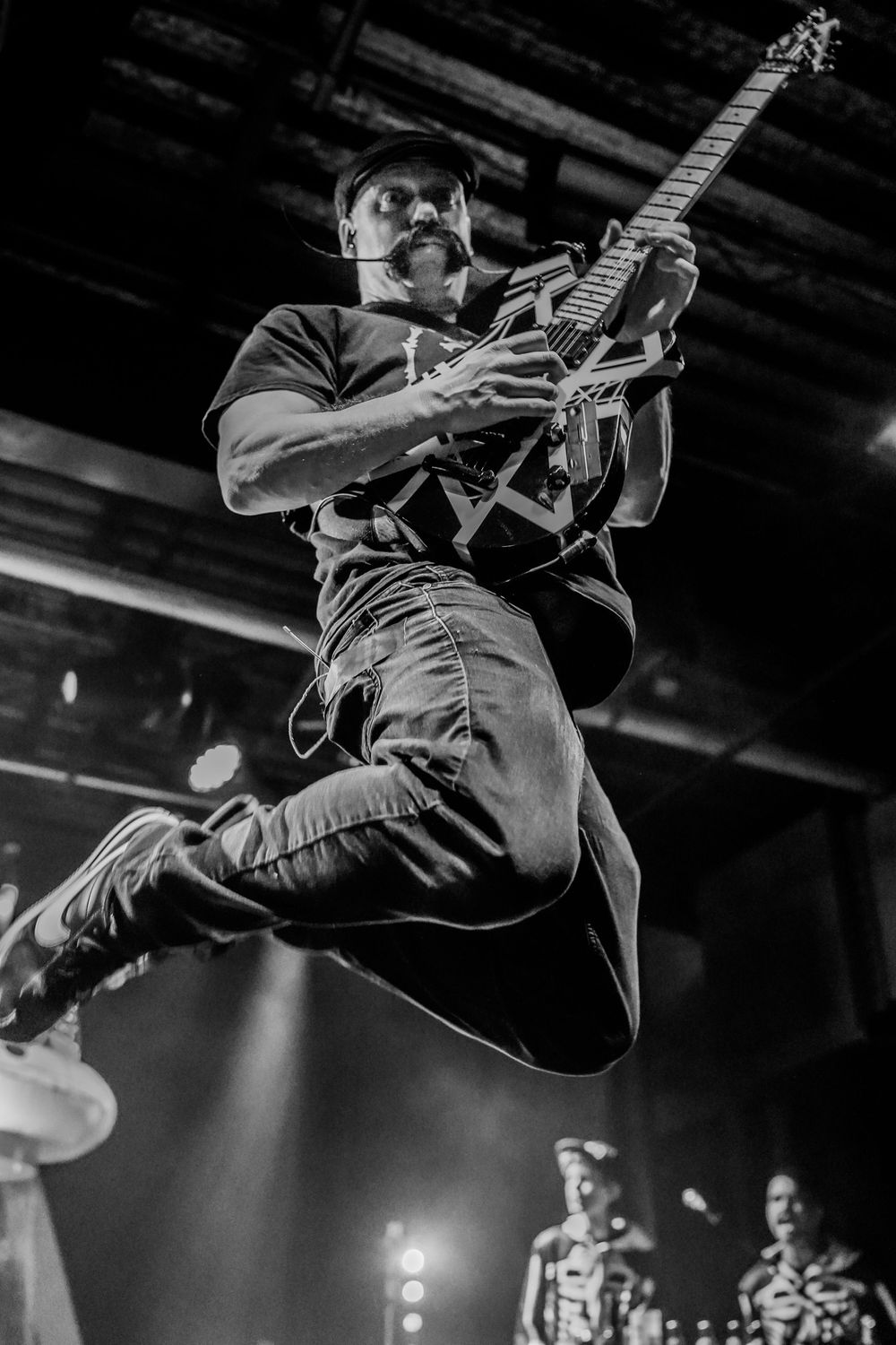 Dynamic black and white photo of a musician leaping high while playing guitar during an energetic stage performance.