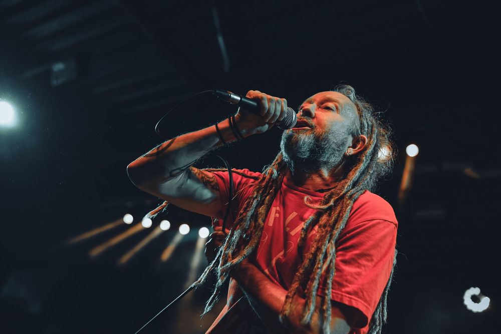 A performer in a red shirt sings passionately into a microphone on a dimly lit concert stage.