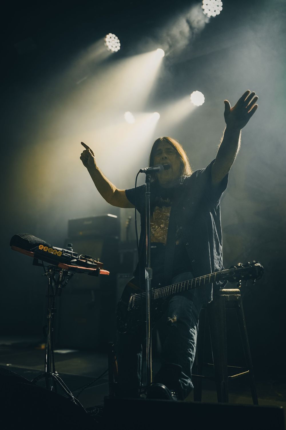 Atmospheric concert scene with performer silhouetted against dramatic spotlights and hazy stage lighting.