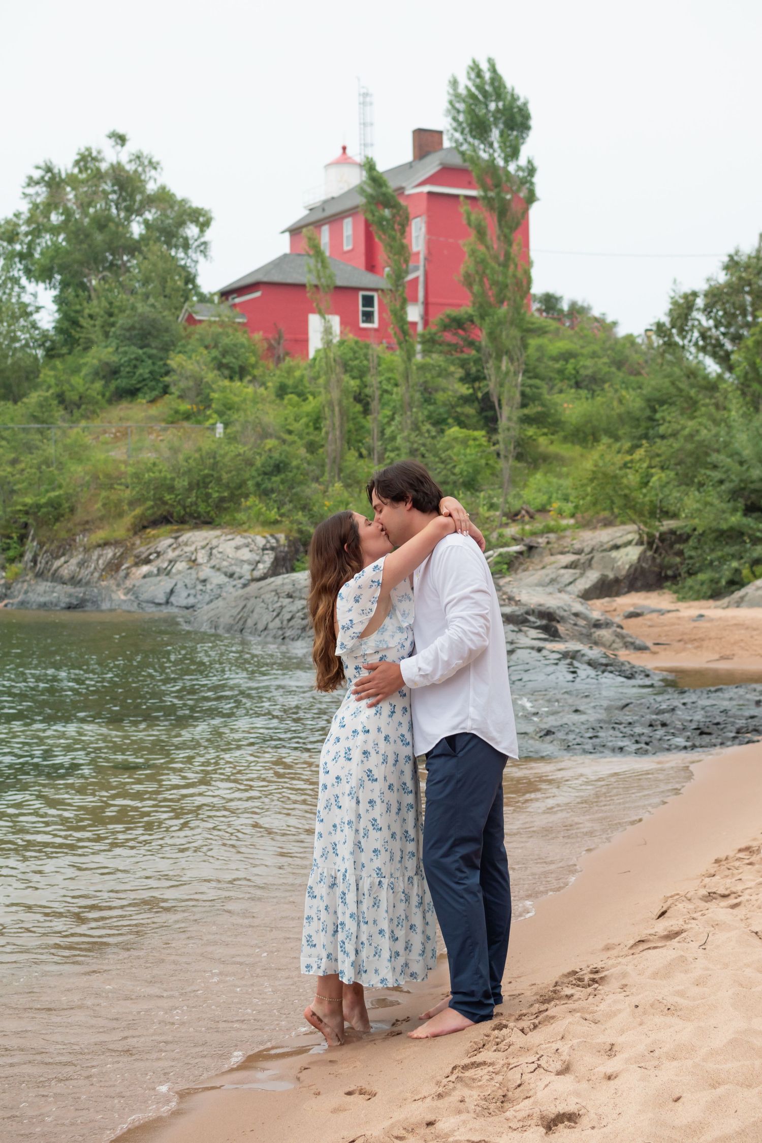 A couple shares a romantic moment on a sandy beach with a red lighthouse and trees in the background.