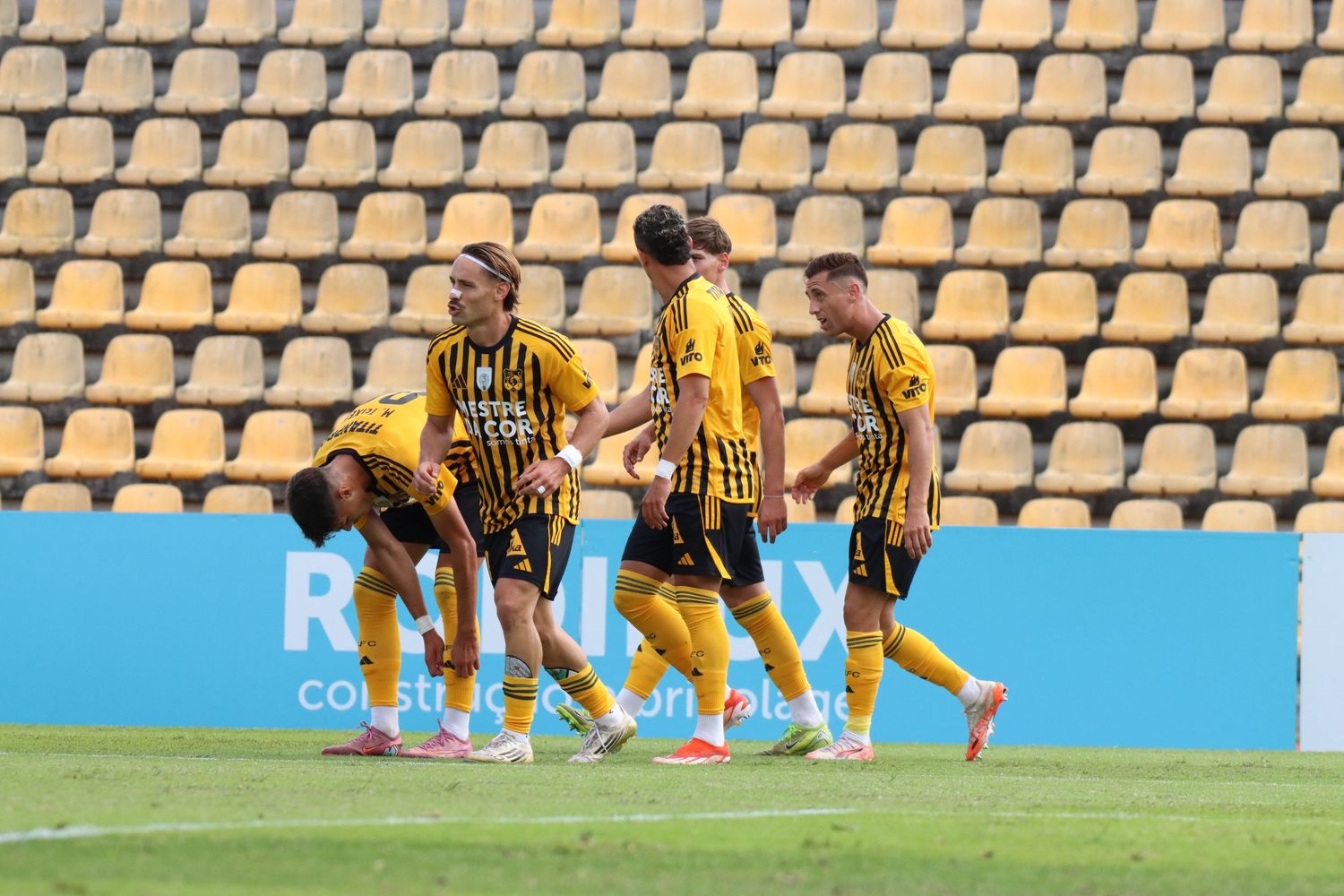 Soccer match in progress at stadium with yellow seats showing players in yellow and white uniforms competing.