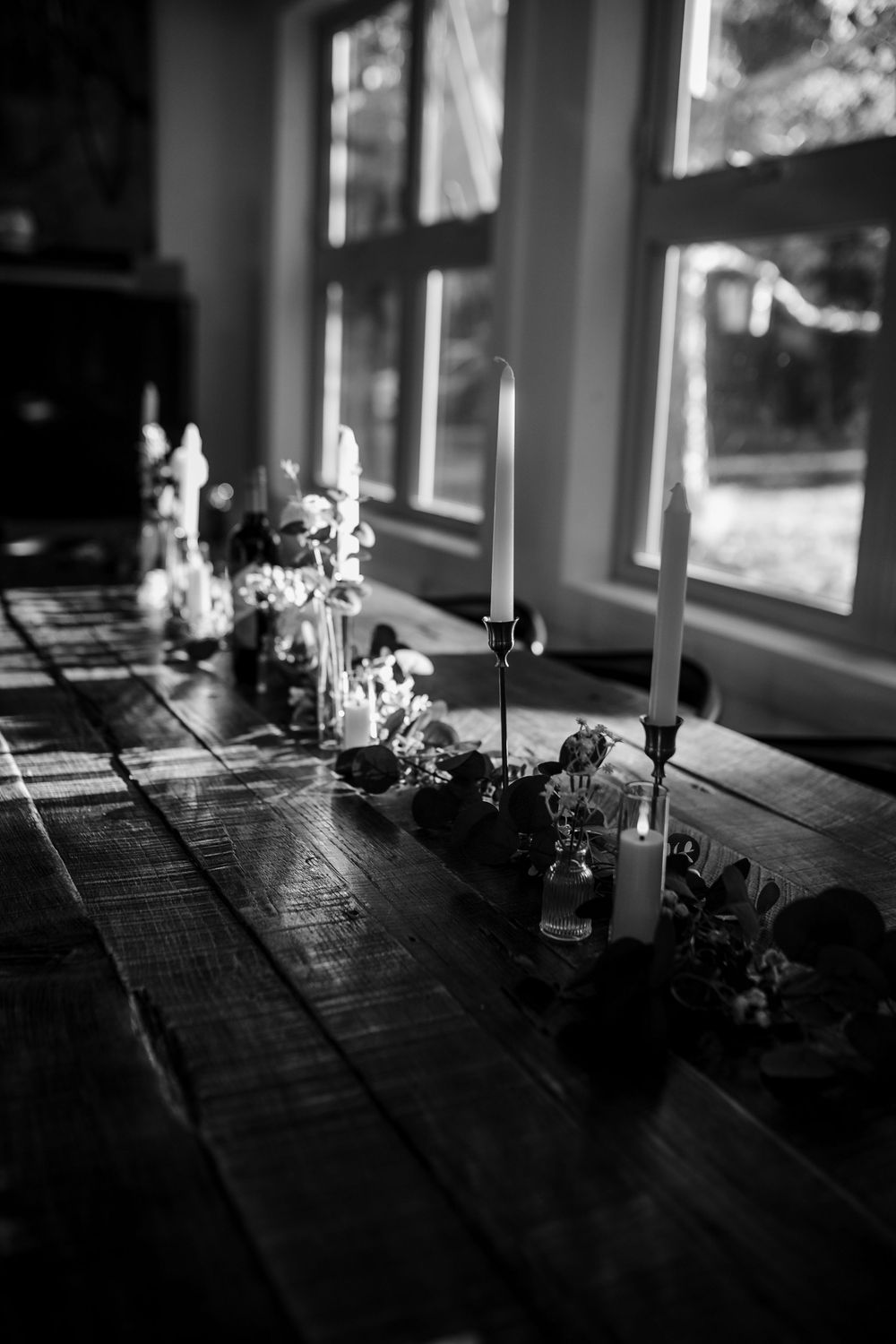 Candlesticks and decorative items line a rustic wooden table by sunlit windows in black and white photography.