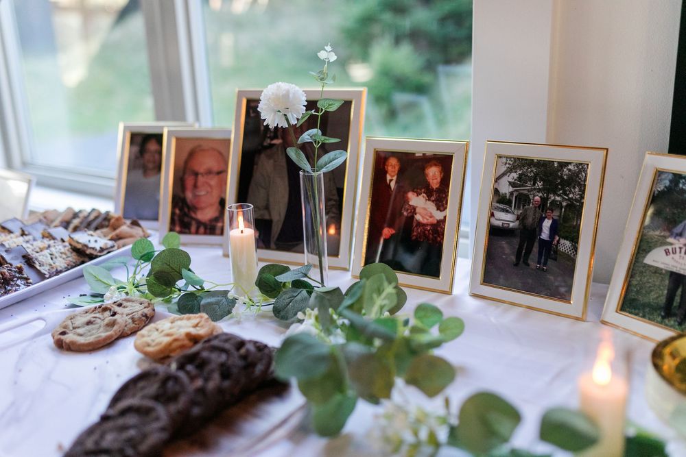 A memorial table display with photographs, candles, and greenery arranged near a window.