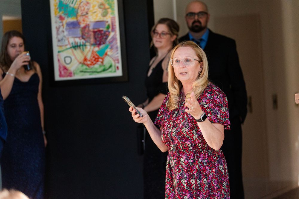 A person in a pink floral dress gestures expressively while speaking in a hallway setting.