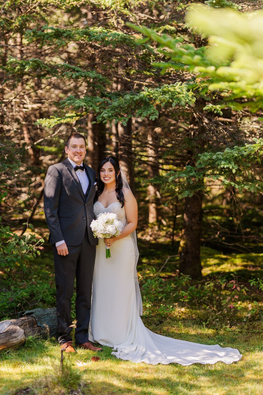 A couple poses for a wedding photo in a wooded setting with the bride wearing a flowing white gown with train.