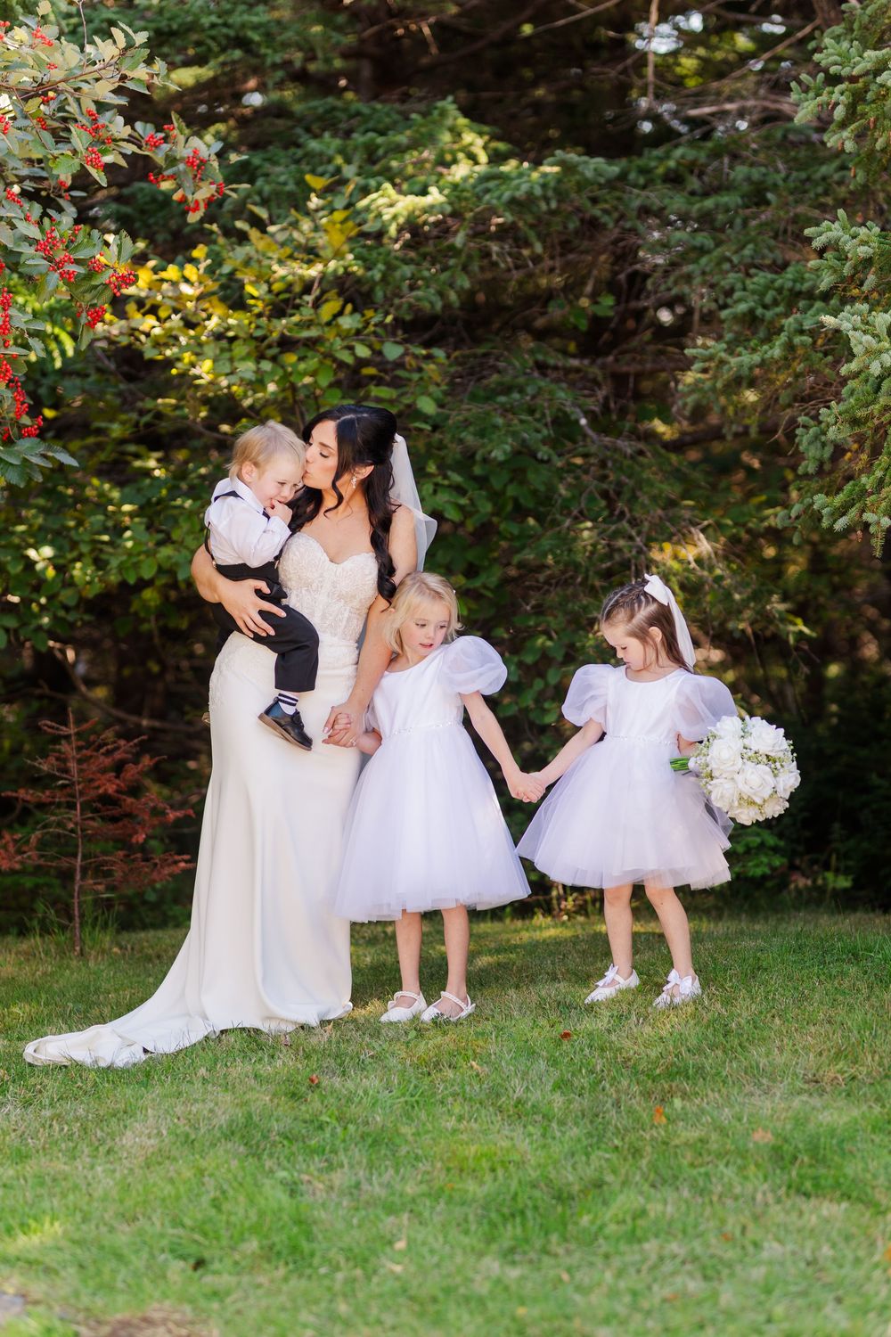 A wedding party with flower girls in white dresses poses together on a grassy lawn surrounded by trees.
