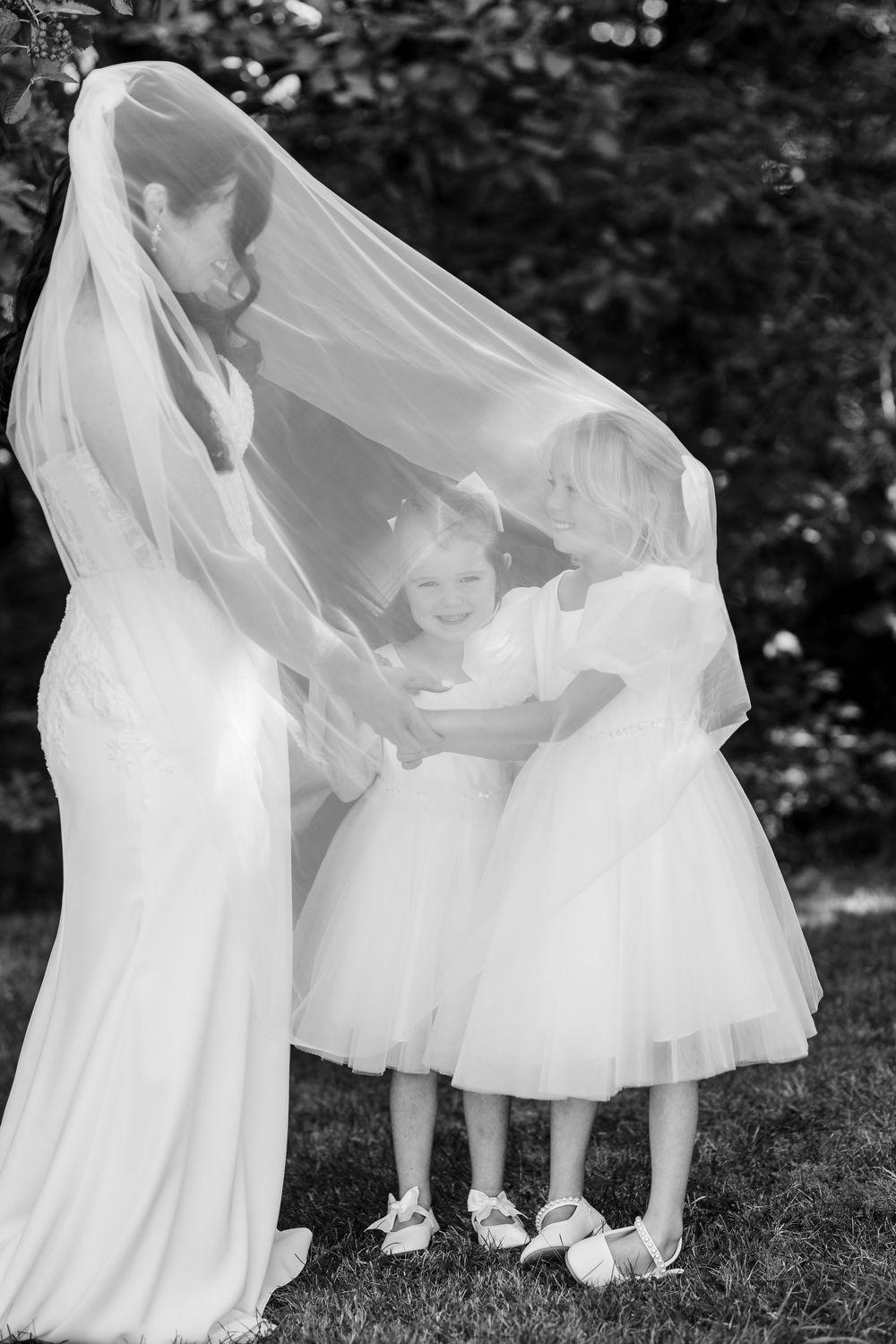 A bride in a flowing white gown shares a playful moment under her veil with a young flower girl in a tulle dress.
