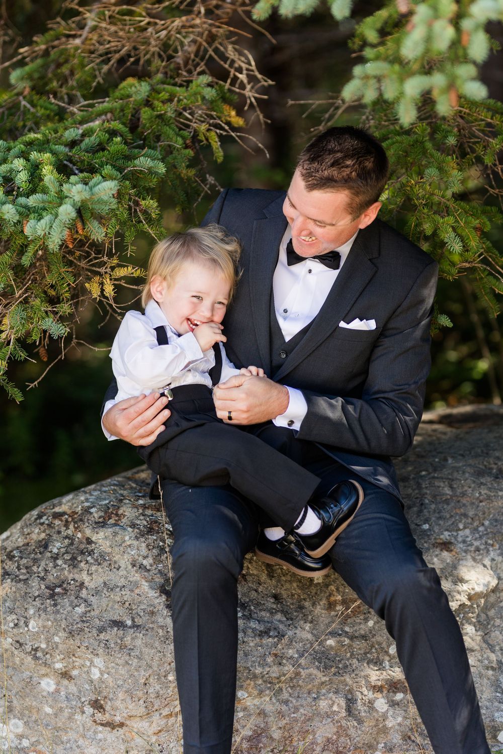A formal-dressed pair share a joyful moment while sitting on a large rock under evergreen branches.