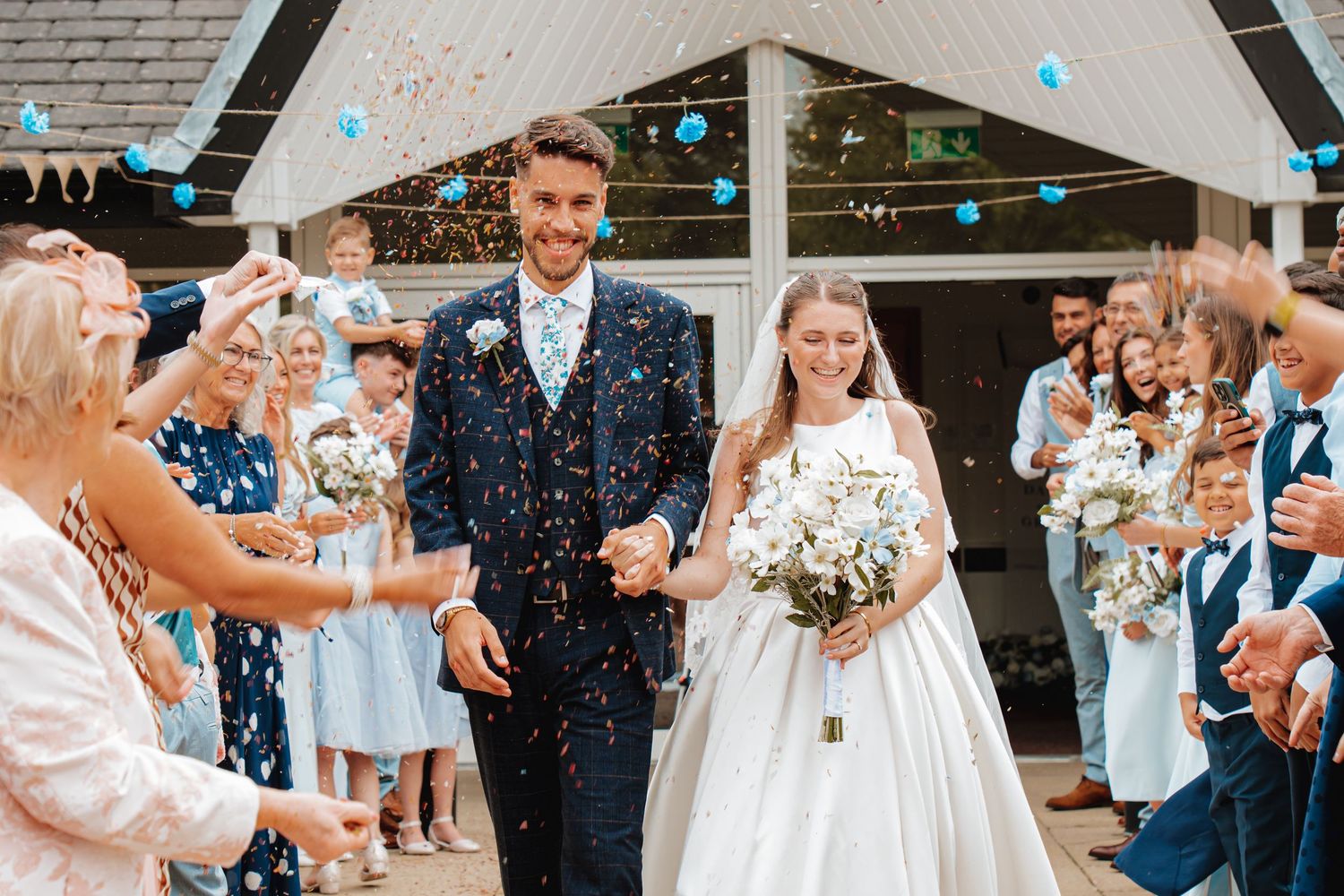 Bride and groom walking through confetti shower surrounded by cheering wedding guests outside rustic venue.