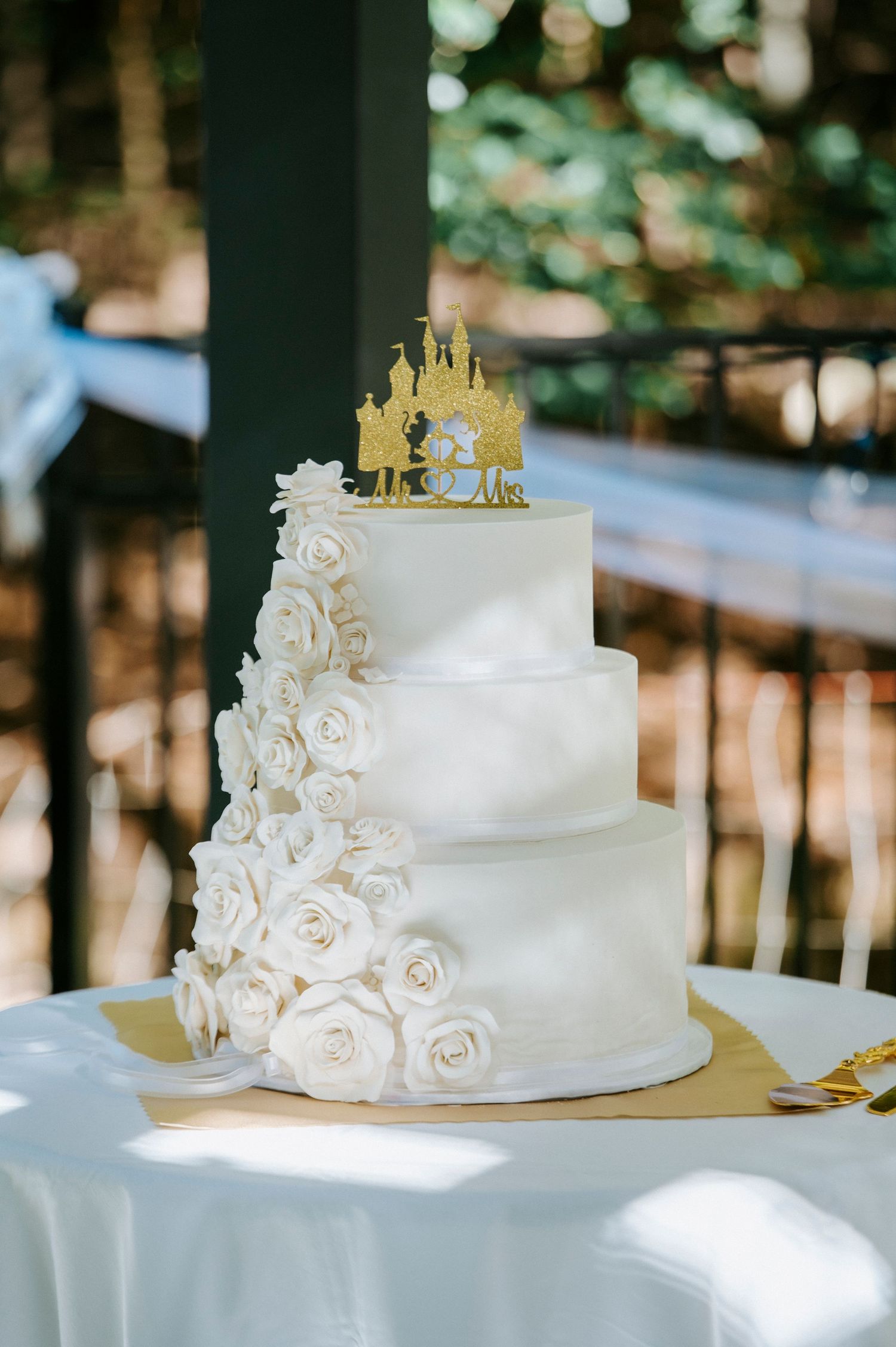 Elegant white wedding cake with cascading sugar roses and a golden castle topper displayed outdoors.