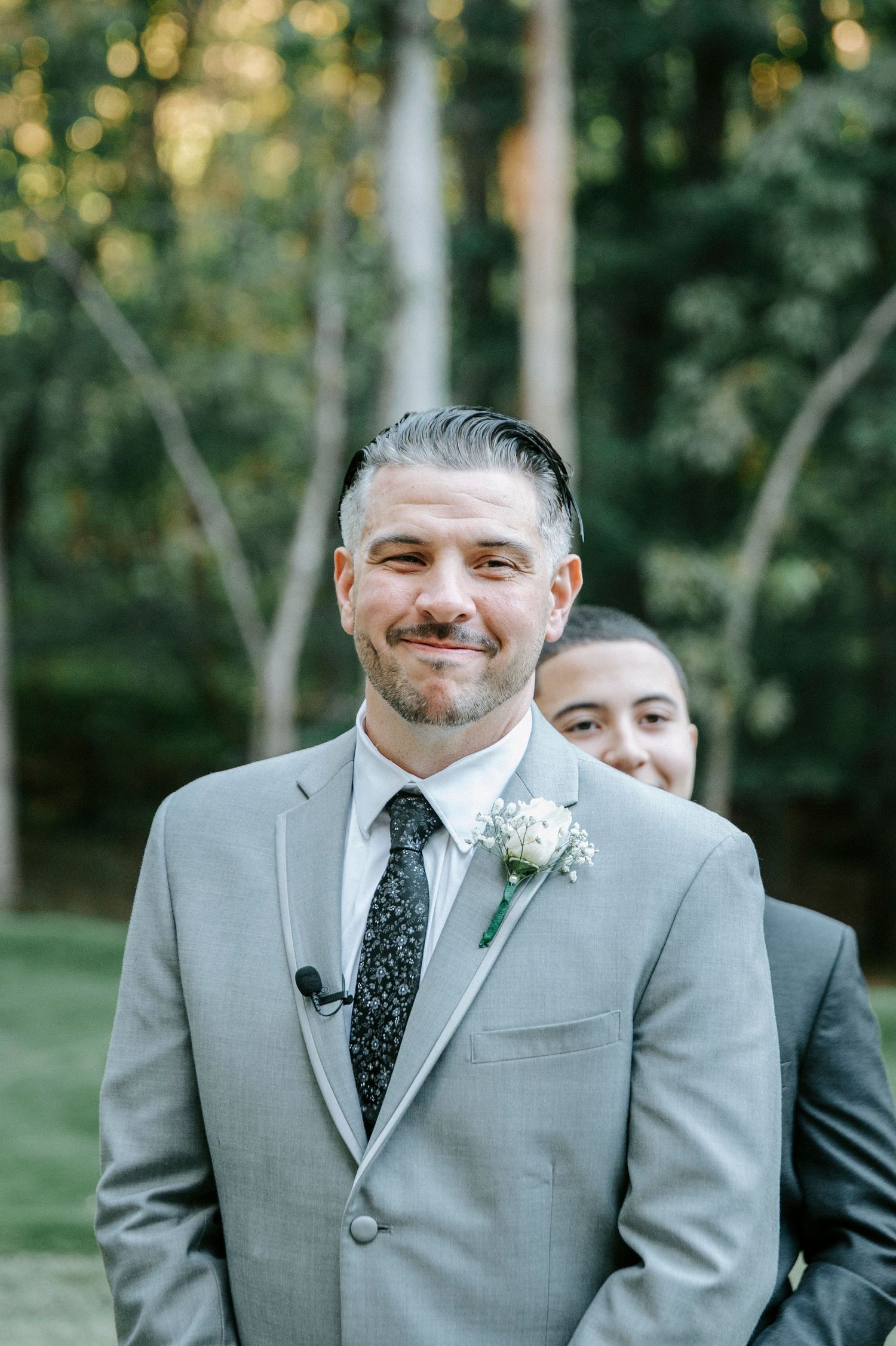 A wedding party member in a light gray suit with a polka dot tie and boutonniere smiles outdoors.