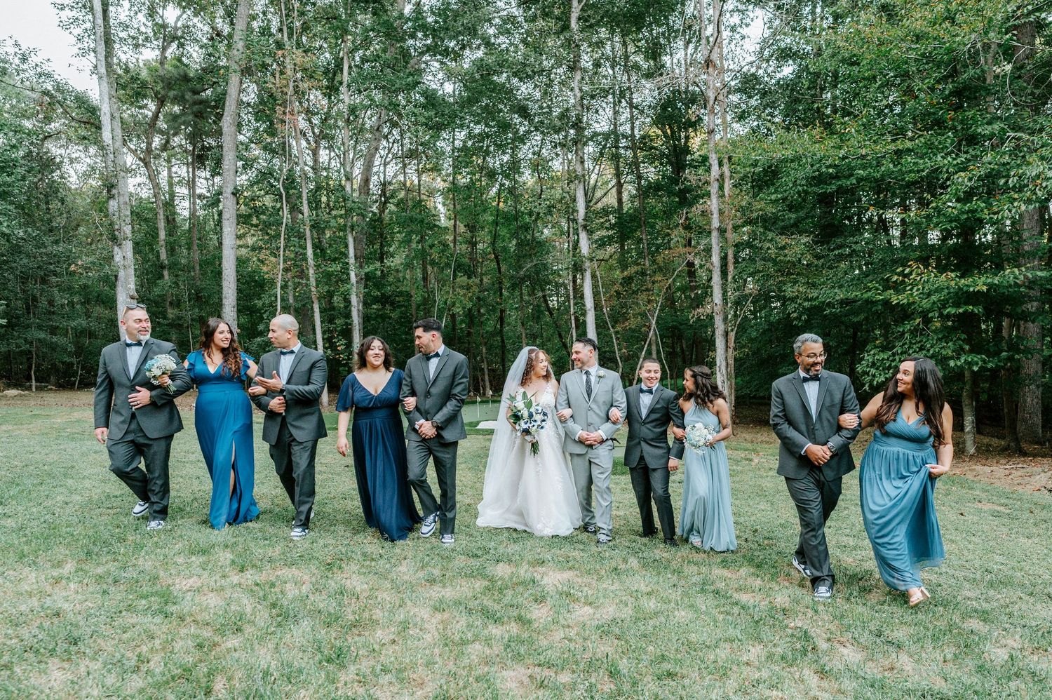 Wedding party in formal attire poses together against a backdrop of lush green trees and foliage.
