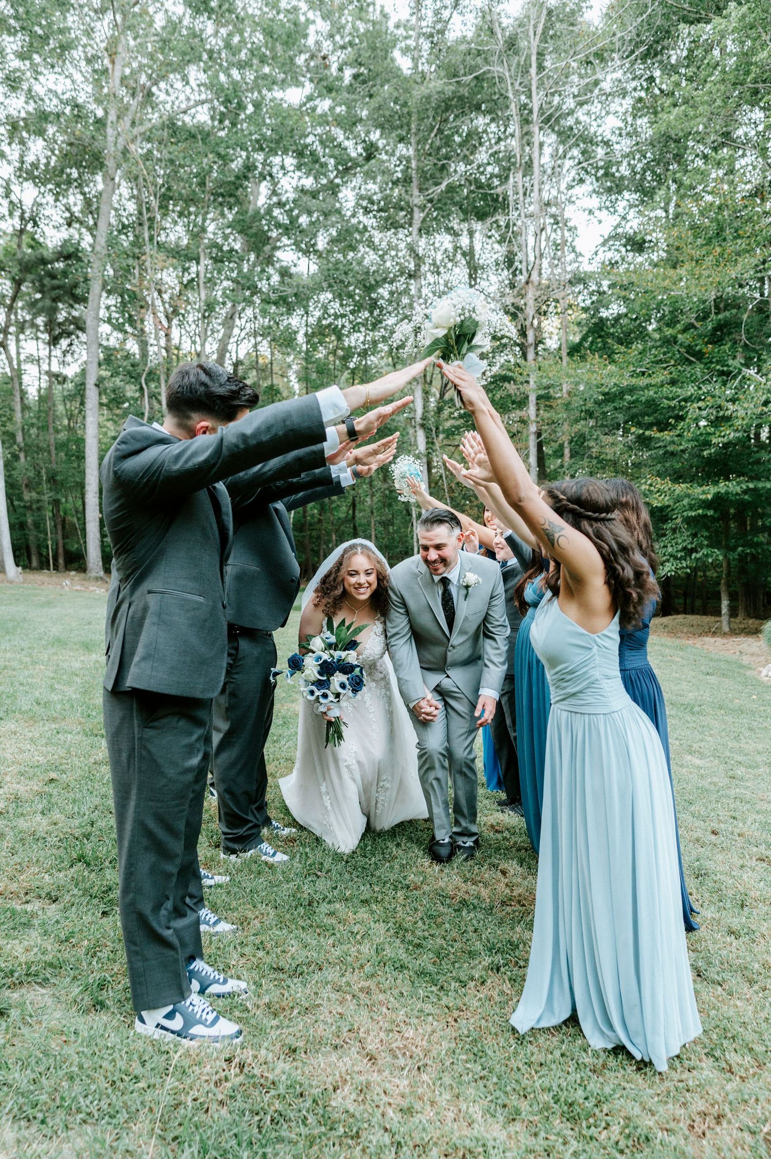 Wedding party in light blue dresses and dark suits performs a fun dance move together in an outdoor garden setting.