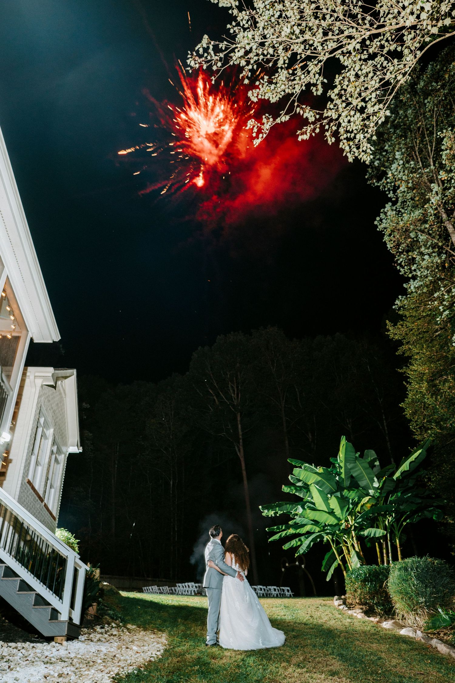 A romantic wedding scene with fireworks lighting up the night sky over a white building and lush garden.
