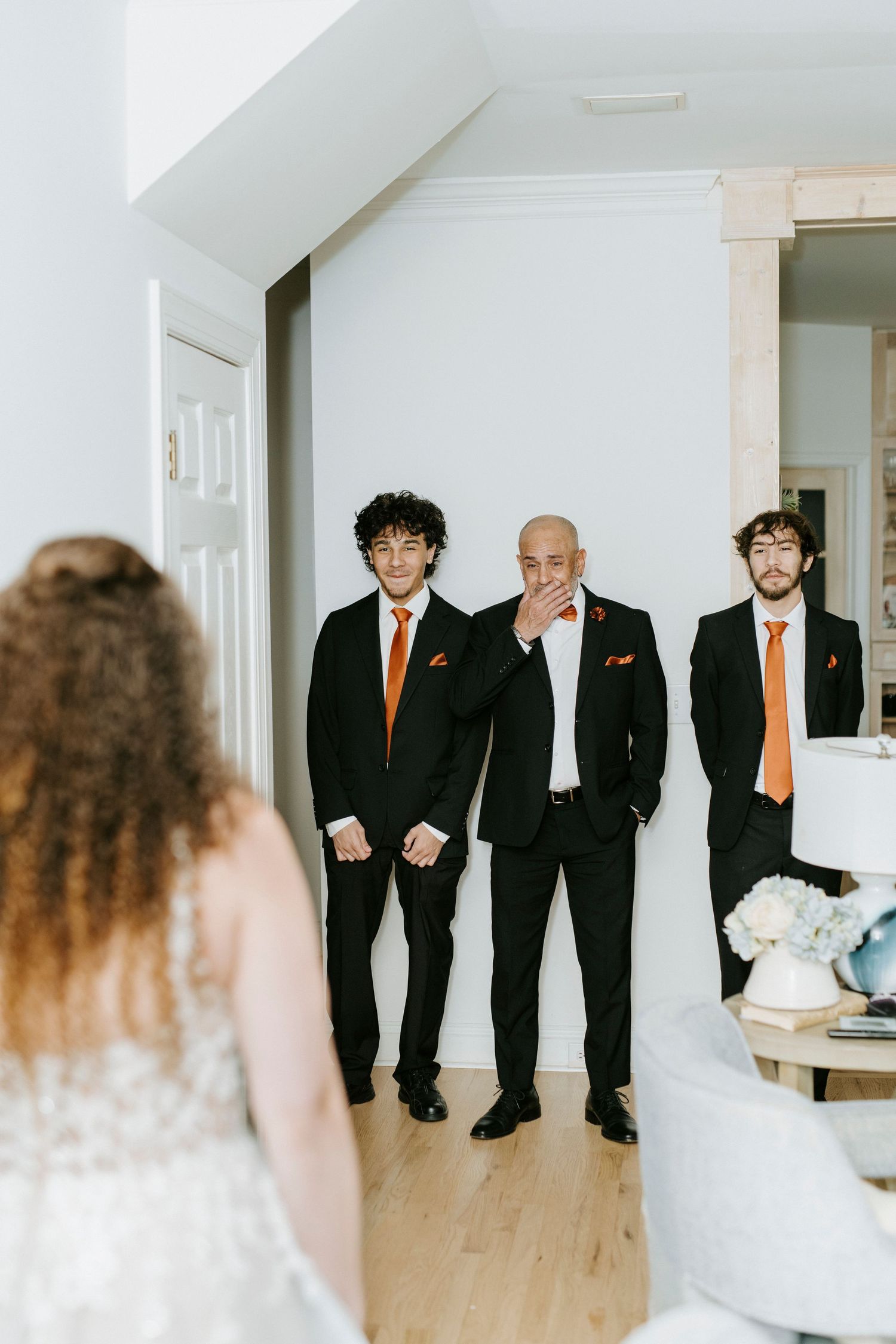 Groomsmen in black suits with orange ties lined up in a bright white room.