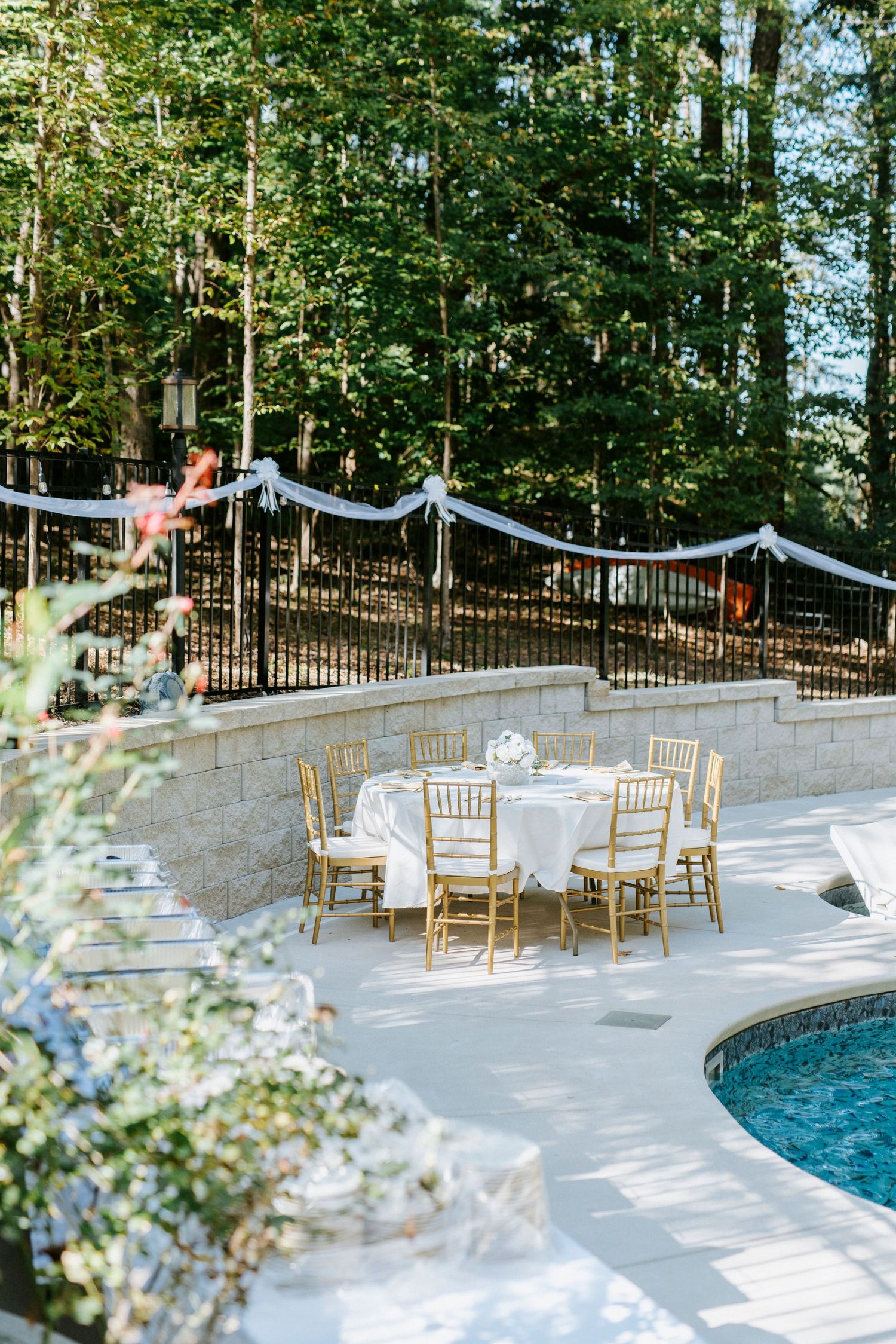 Elegant poolside wedding reception setup with white chairs and tables surrounded by lush greenery.
