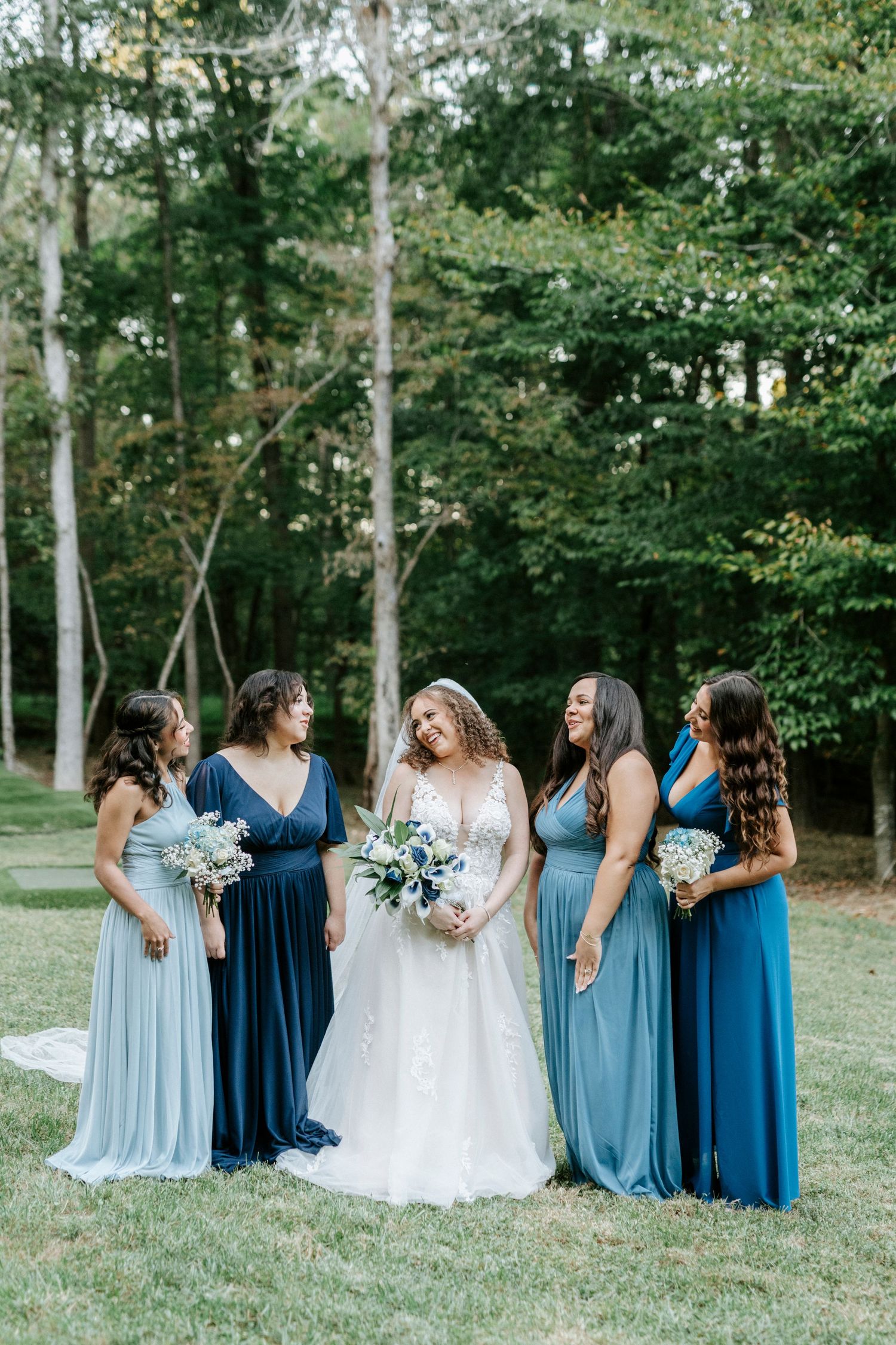 Wedding party in coordinated blue dresses standing together on a grassy lawn with bouquets.