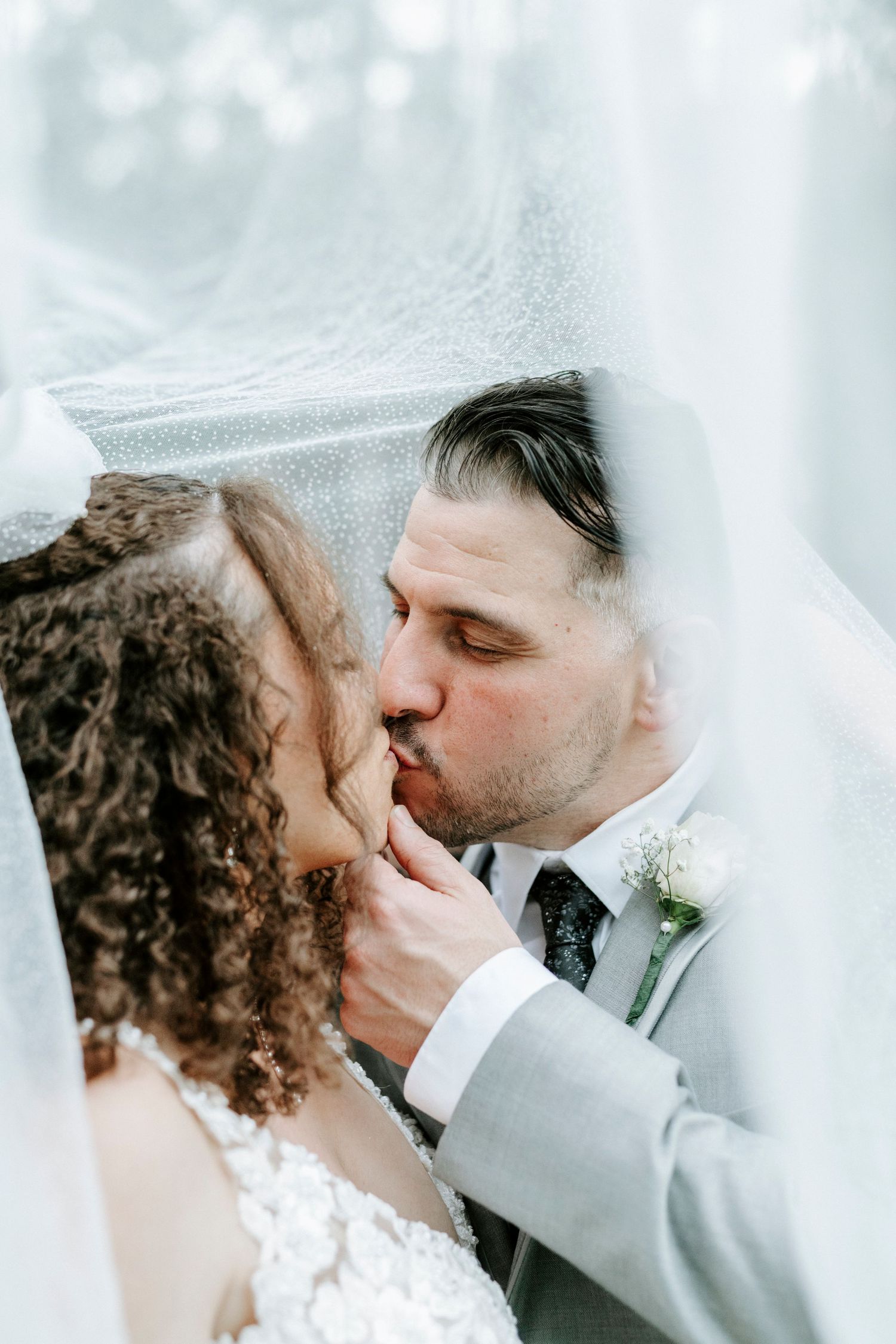 A tender wedding moment captured under a white veil as newlyweds share their first kiss in soft lighting.