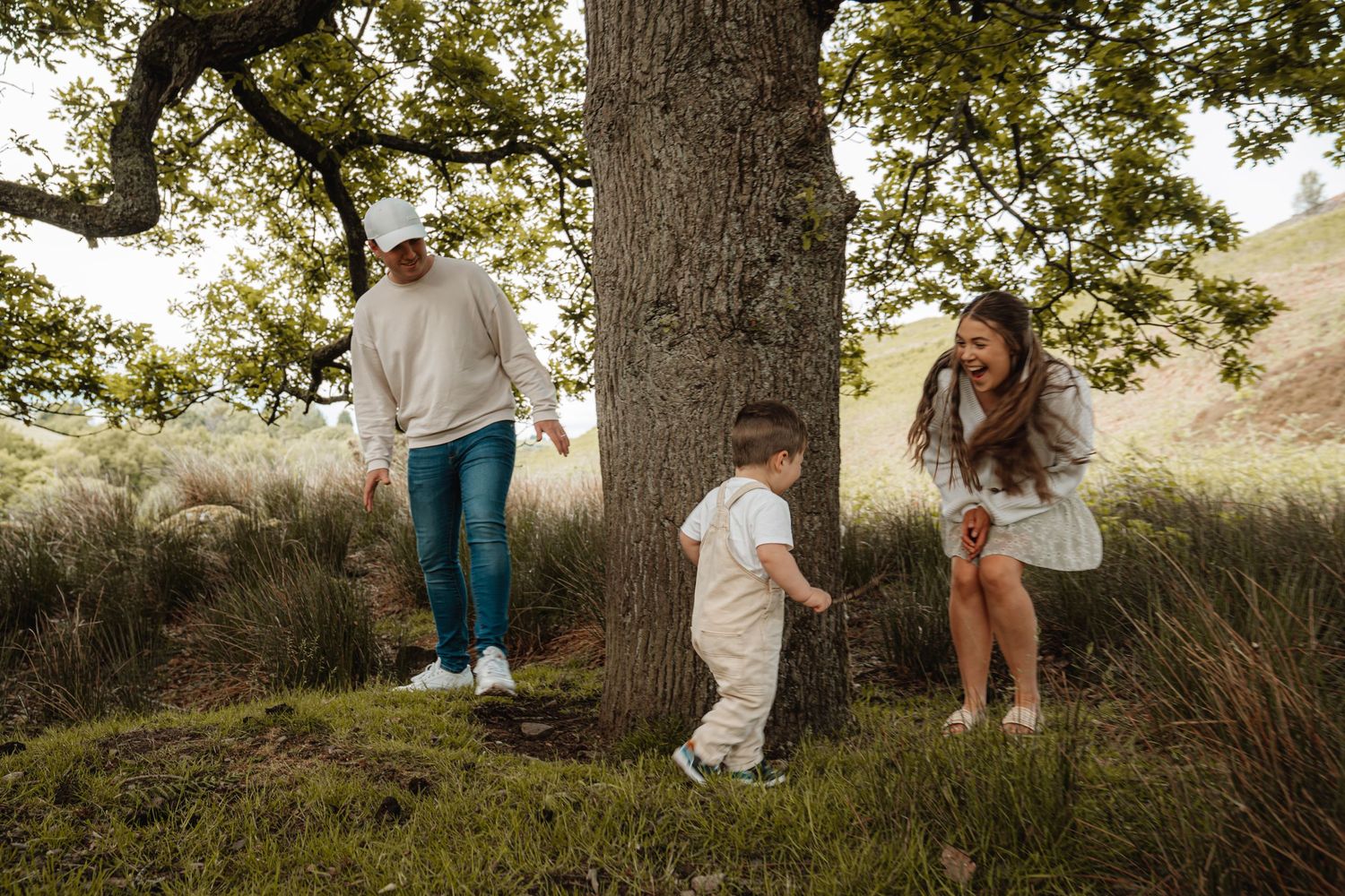 Young family with toddler playing around a large oak tree in a scenic countryside landscape.