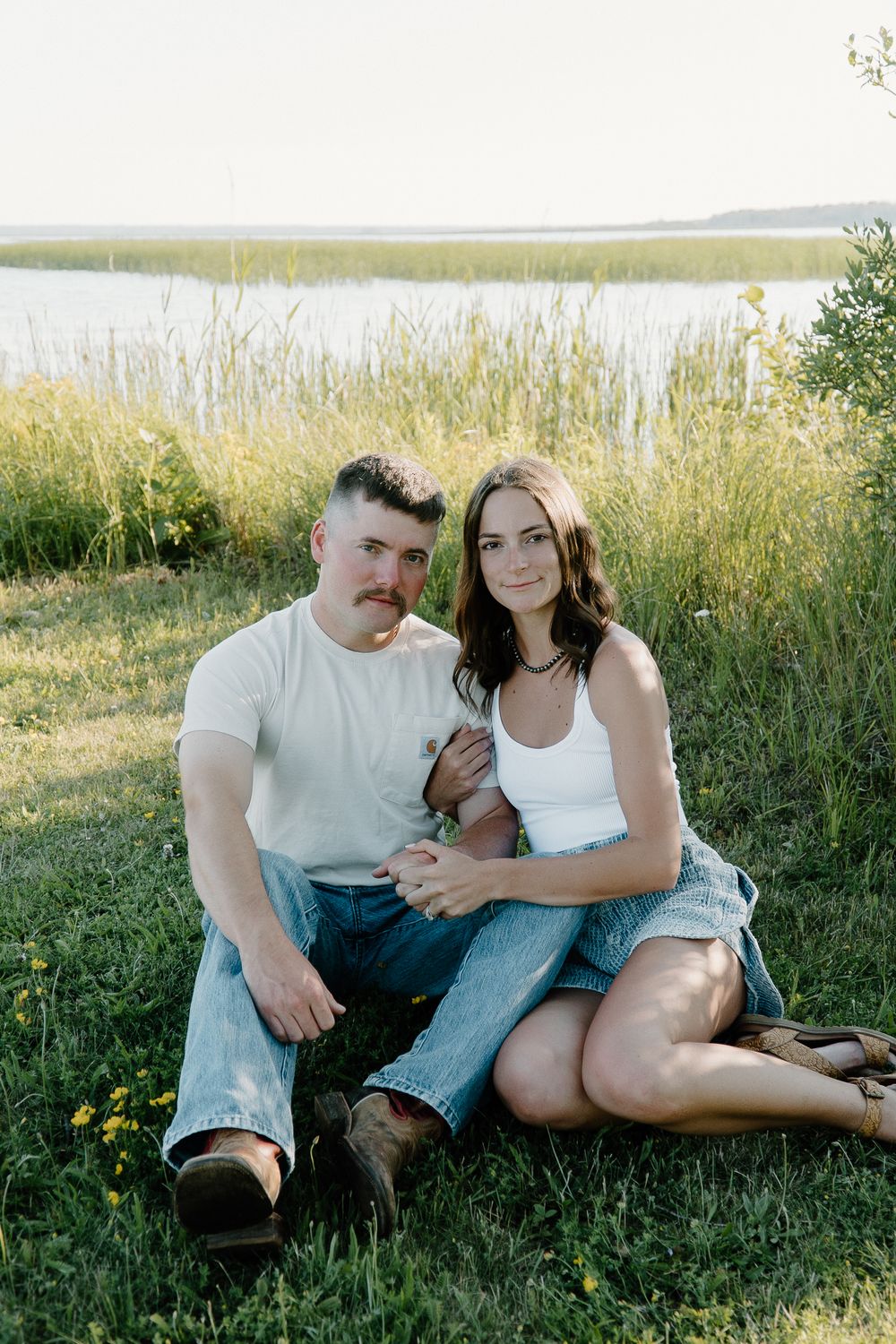A couple sits together on grassy ground during a romantic outdoor photoshoot at golden hour.