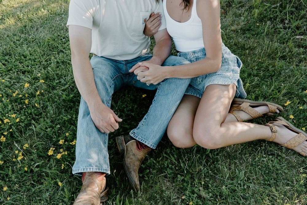 A couple sits together on green grass in a romantic outdoor setting wearing casual denim and white clothing.