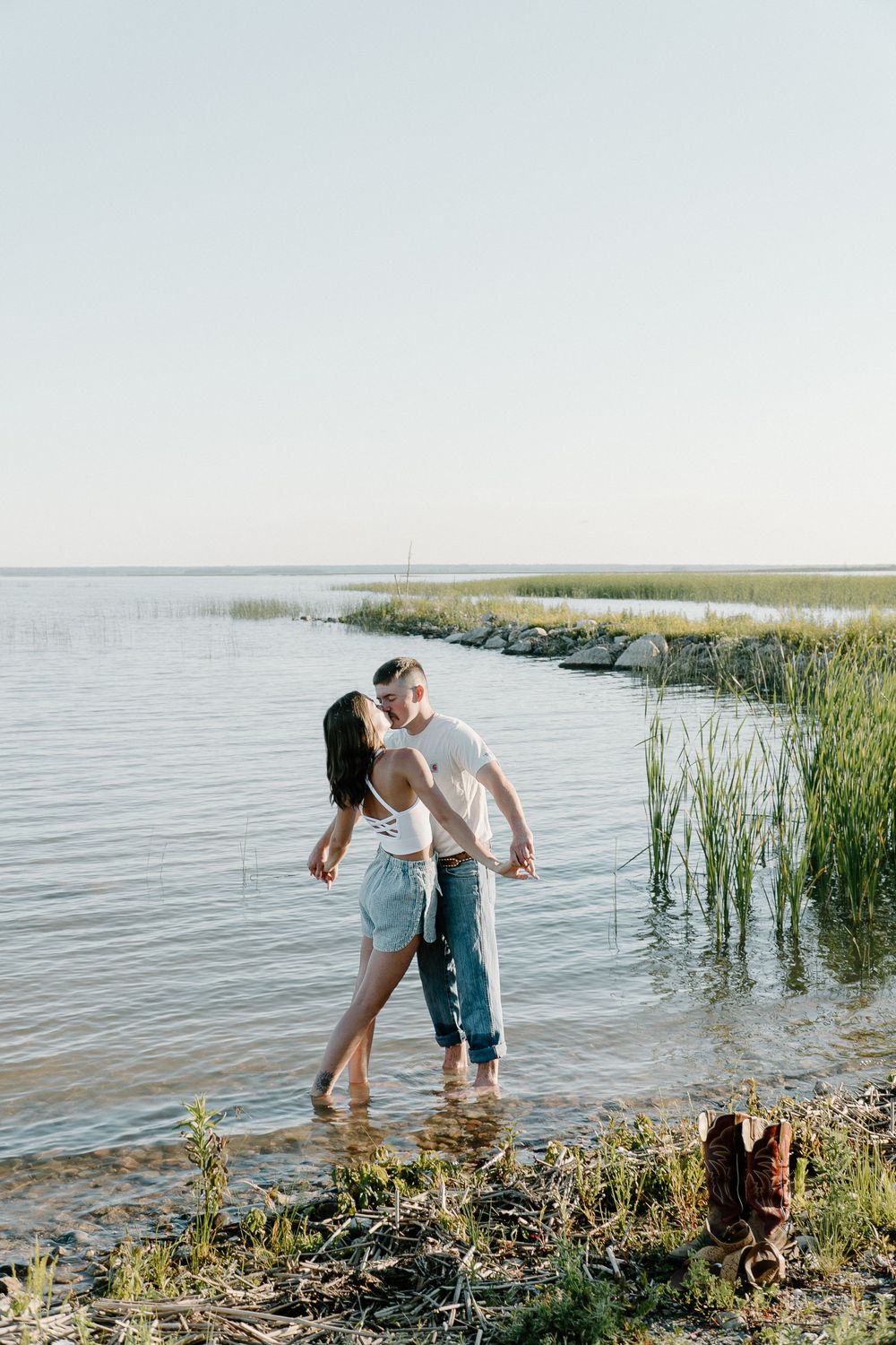 A couple shares a romantic moment by the water's edge with tall grass and reeds in the background.
