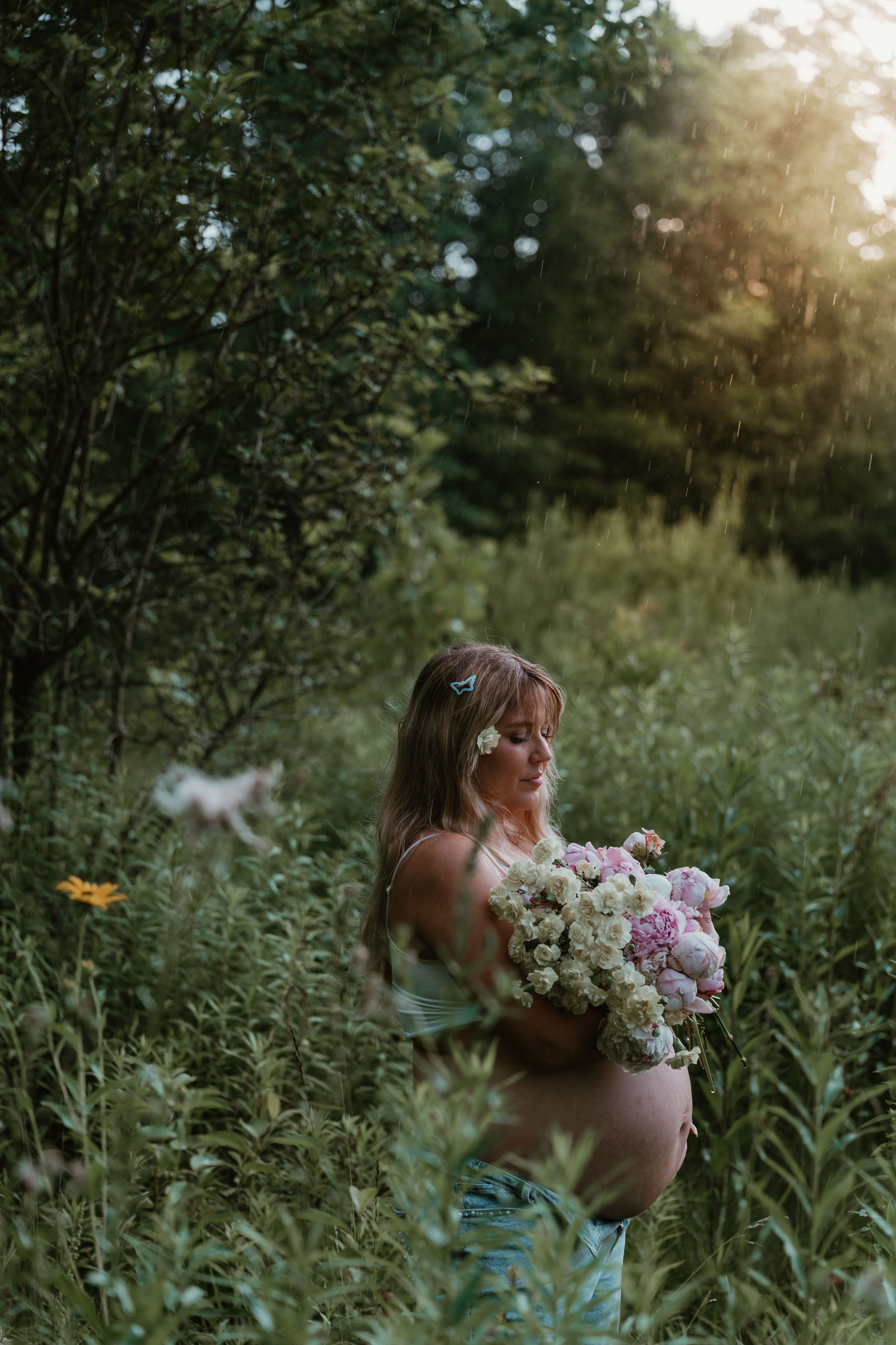 Pregnant woman holding floral bouquet in sunlit green forest clearing at dusk.