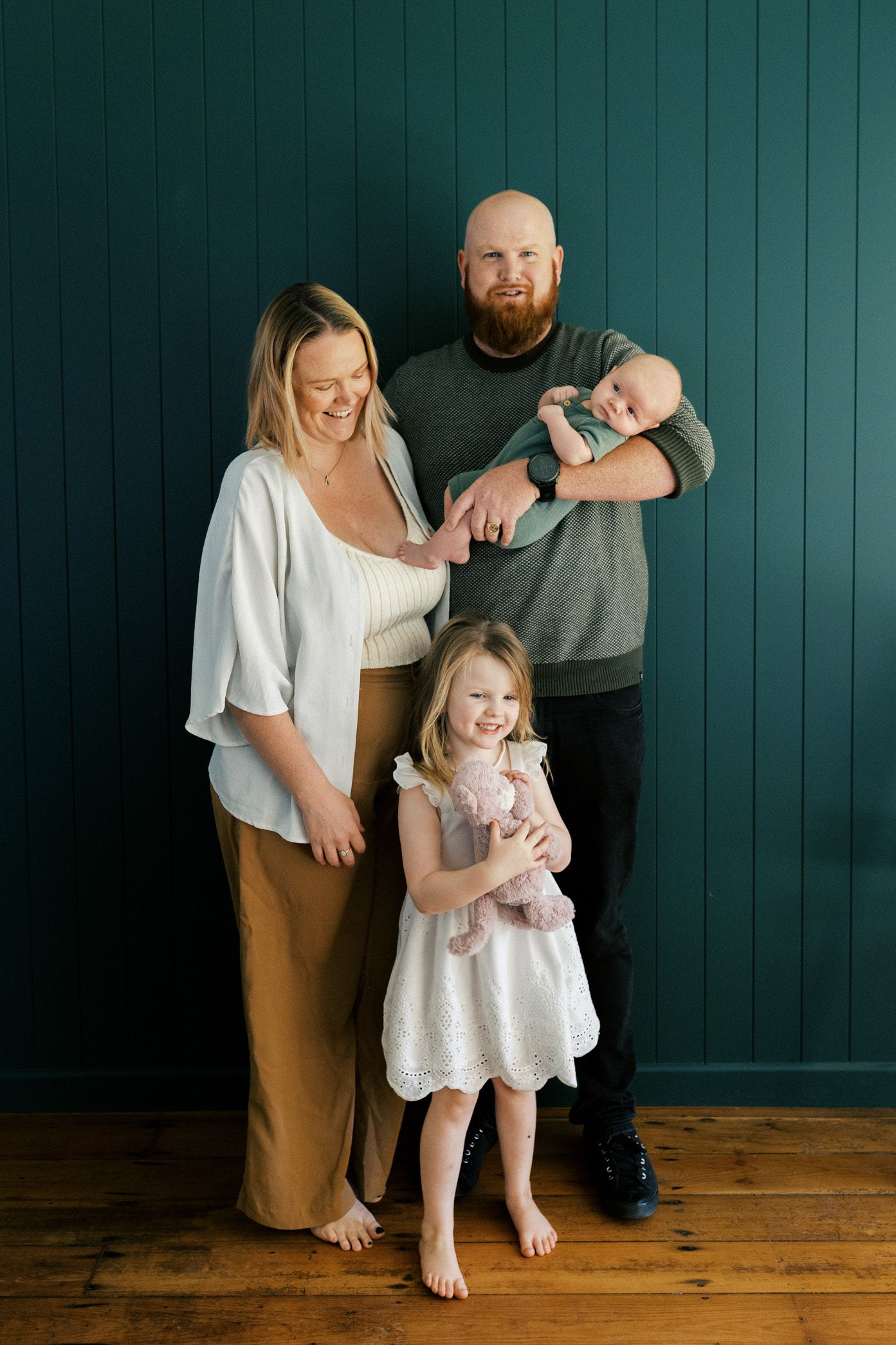 A family in coordinated neutral outfits poses against a dark teal wall and wooden floor in a lifestyle portrait series.