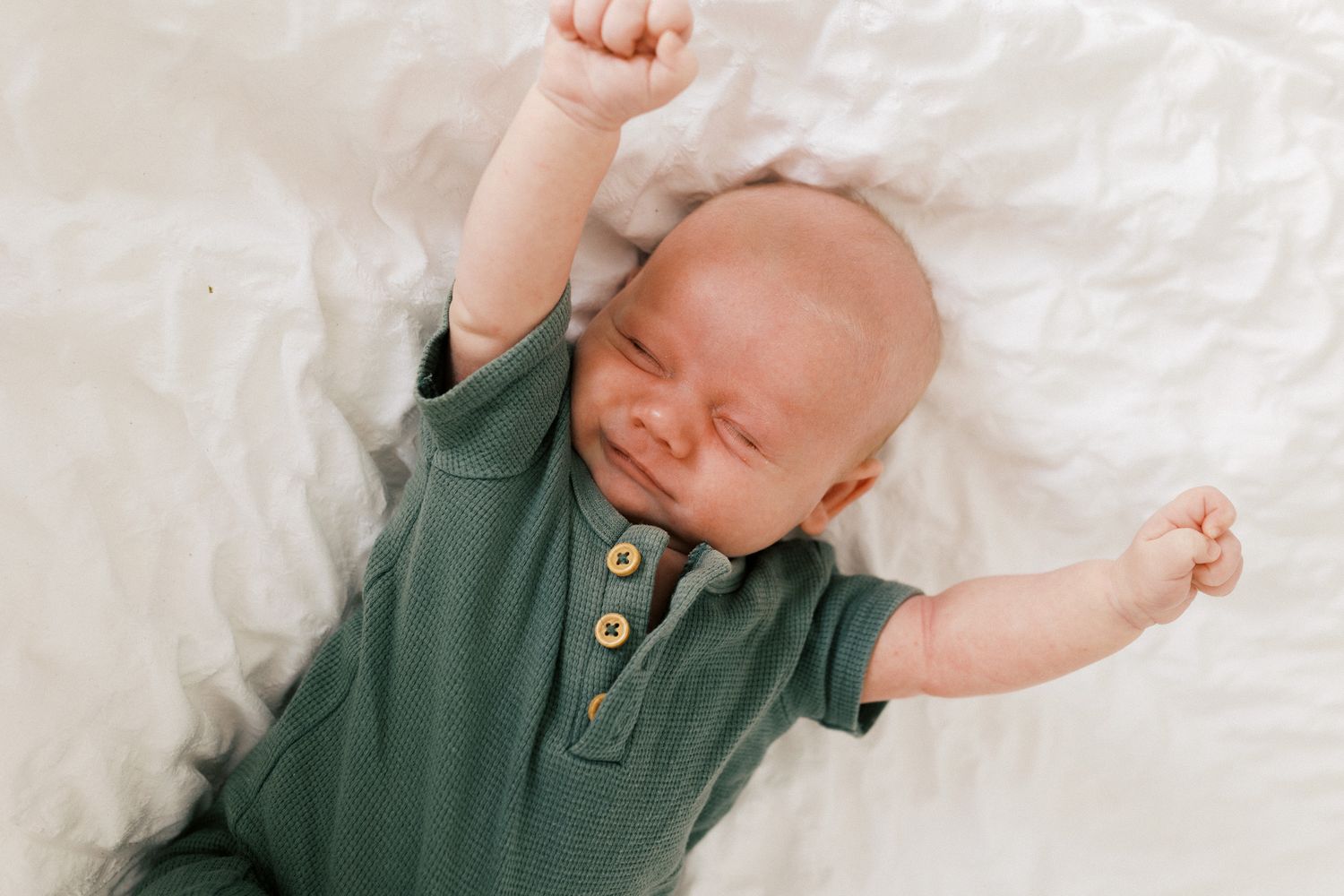 Baby wearing green onesie lying on white blanket stretches arms overhead during playtime.