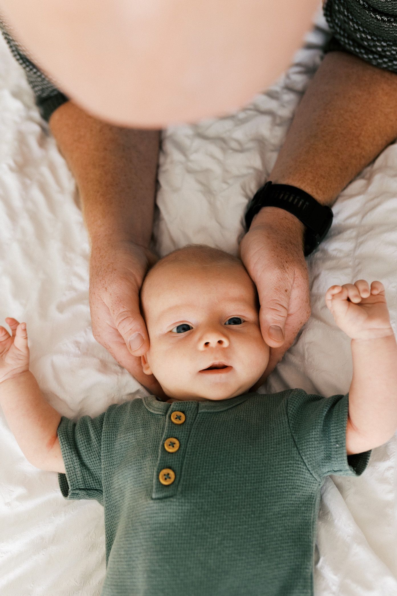 Hands gently hold a small baby wearing a green knit top while lying on white bedding.