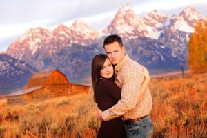 A couple embraces in front of rustic barn and snow-capped Teton mountains during golden autumn sunset.