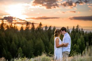 Couple embracing at sunset with evergreen forest backdrop and dramatic sky in a romantic outdoor engagement photo.
