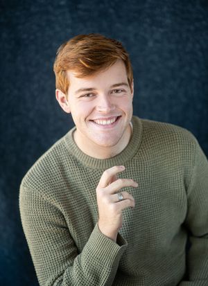 Smiling young man with red hair wearing a green waffle-knit sweater poses against a dark blue background.