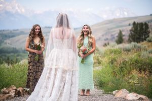 Bride in lace gown with veil stands before two bridesmaids holding bouquets, with mountain scenery behind.