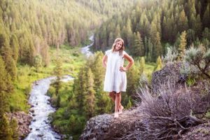 A person in a white dress stands on a rocky overlook above a winding river valley filled with evergreen trees.