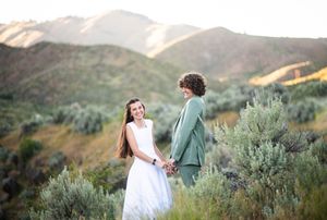 Couple holding hands in a mountain meadow, bride in white dress and groom in green suit, with scenic hills backdrop.
