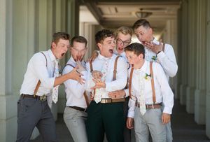 Groomsmen in suspenders and dress shirts laugh together in a sunlit colonnade during a wedding celebration.